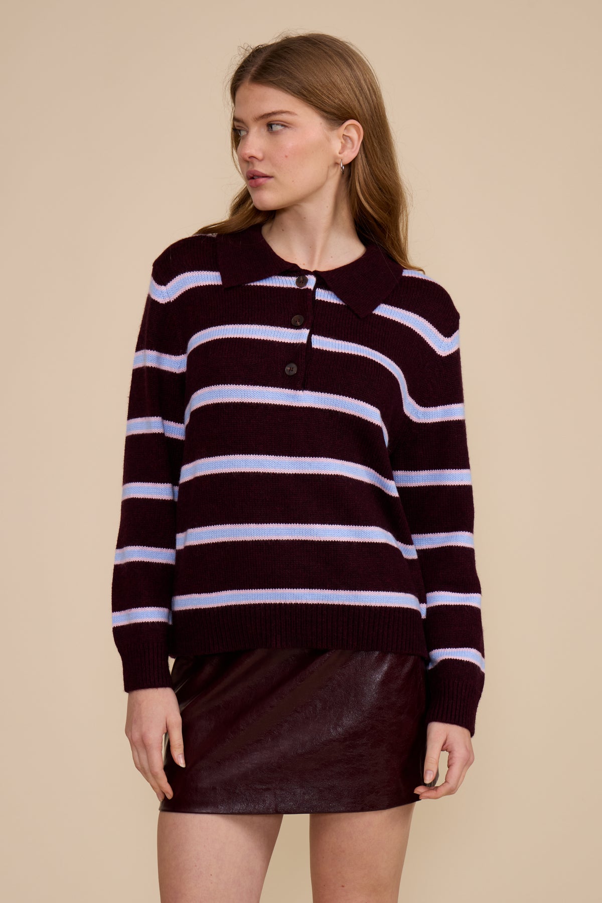 A woman modeling a dark burgundy knit polo sweater with light lavender horizontal stripes, paired with a matching dark burgundy mini skirt, against a solid beige background.