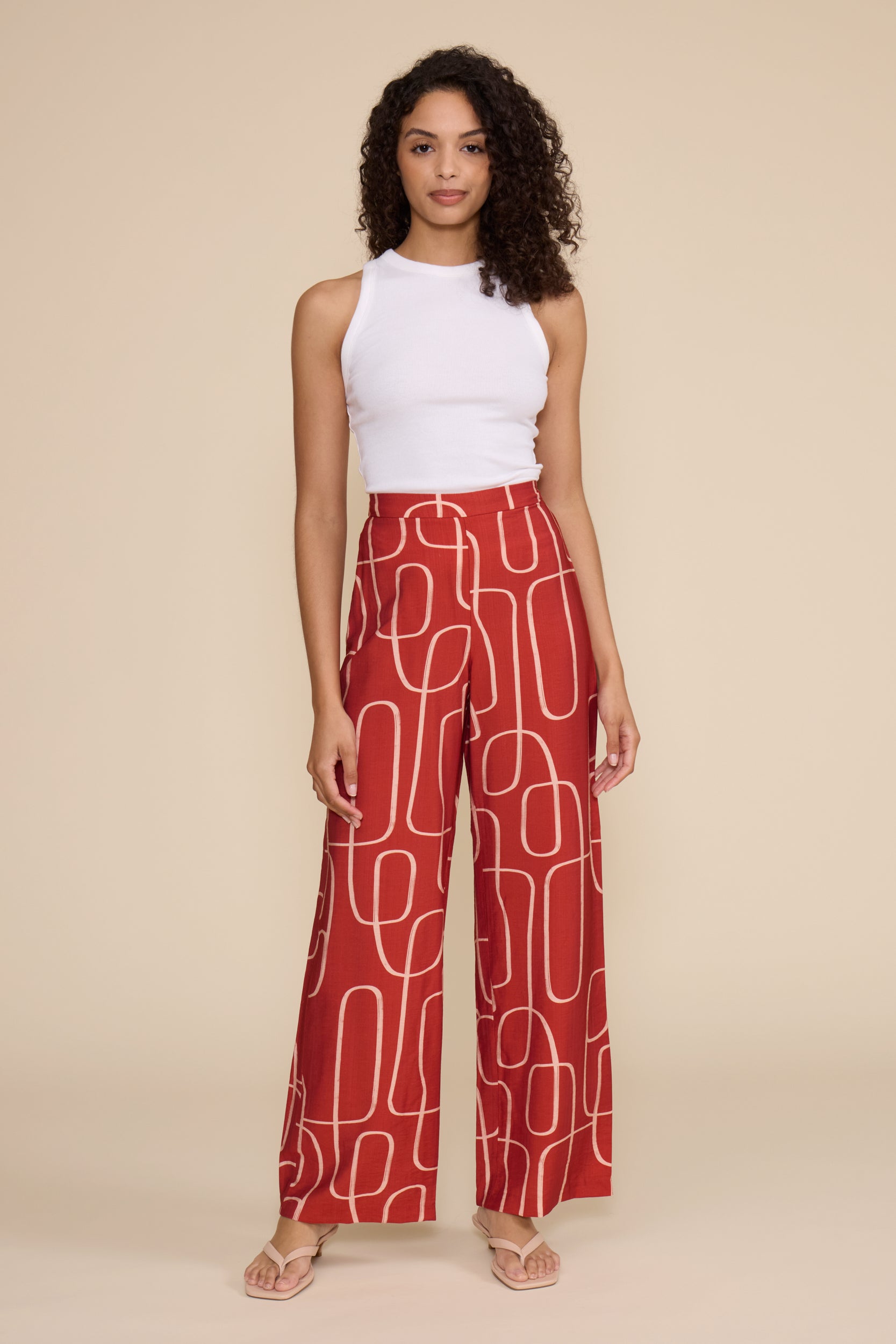 A woman modeling a sleeveless white top and high-waisted red wide-leg trousers with a white abstract line pattern, standing against a solid beige studio background.