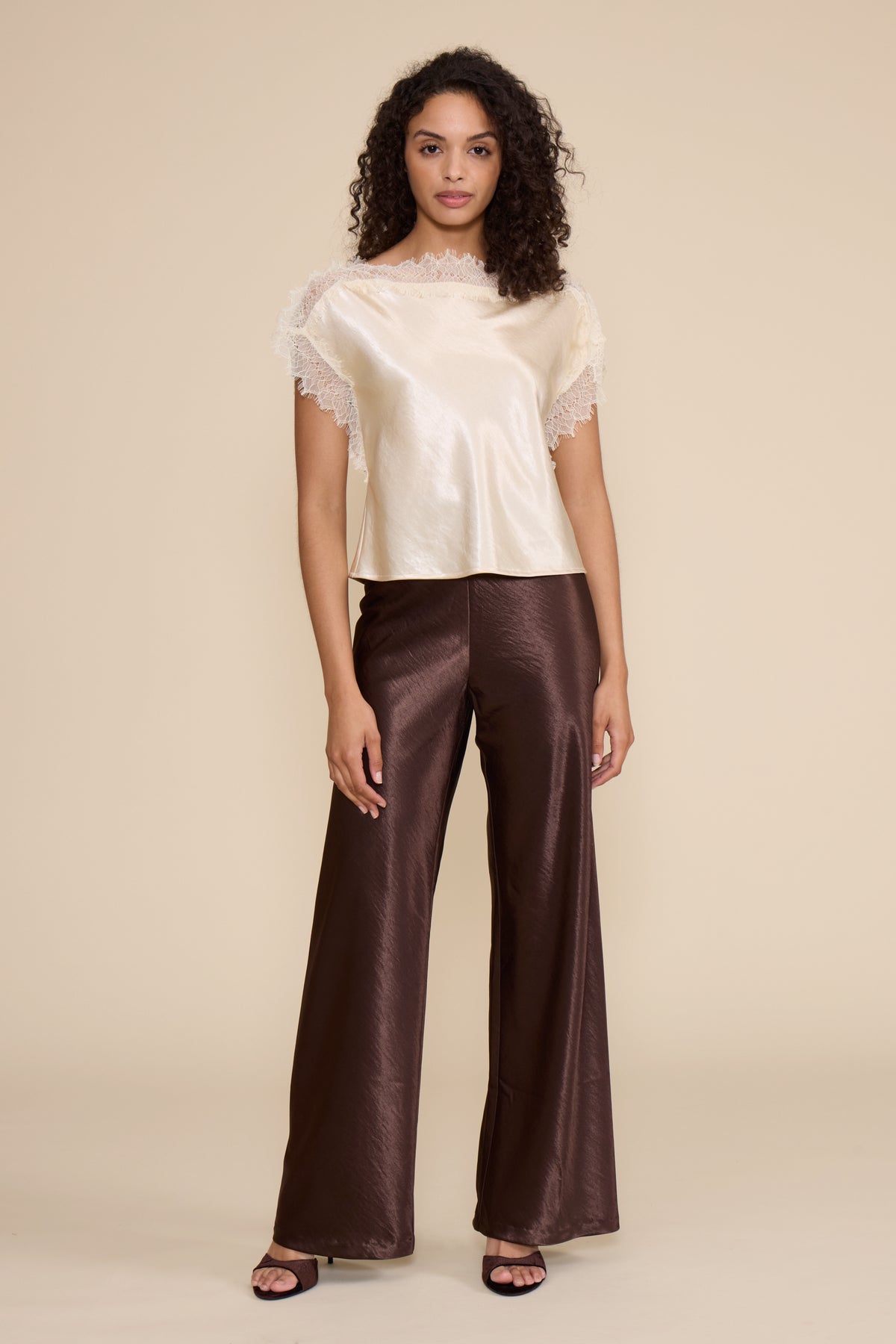 Woman wearing a cream top with lace details and brown pants on a beige background