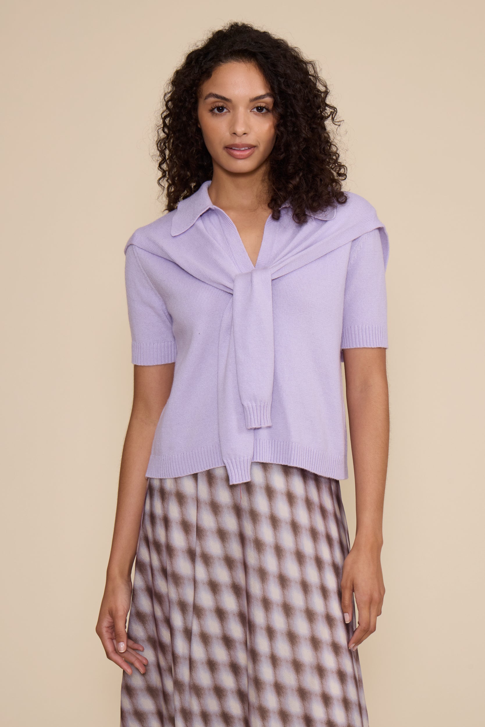 A model wearing a light purple short-sleeve knit top with a wide collar and front tie detail, paired with a brown and white patterned skirt, against a solid beige background.