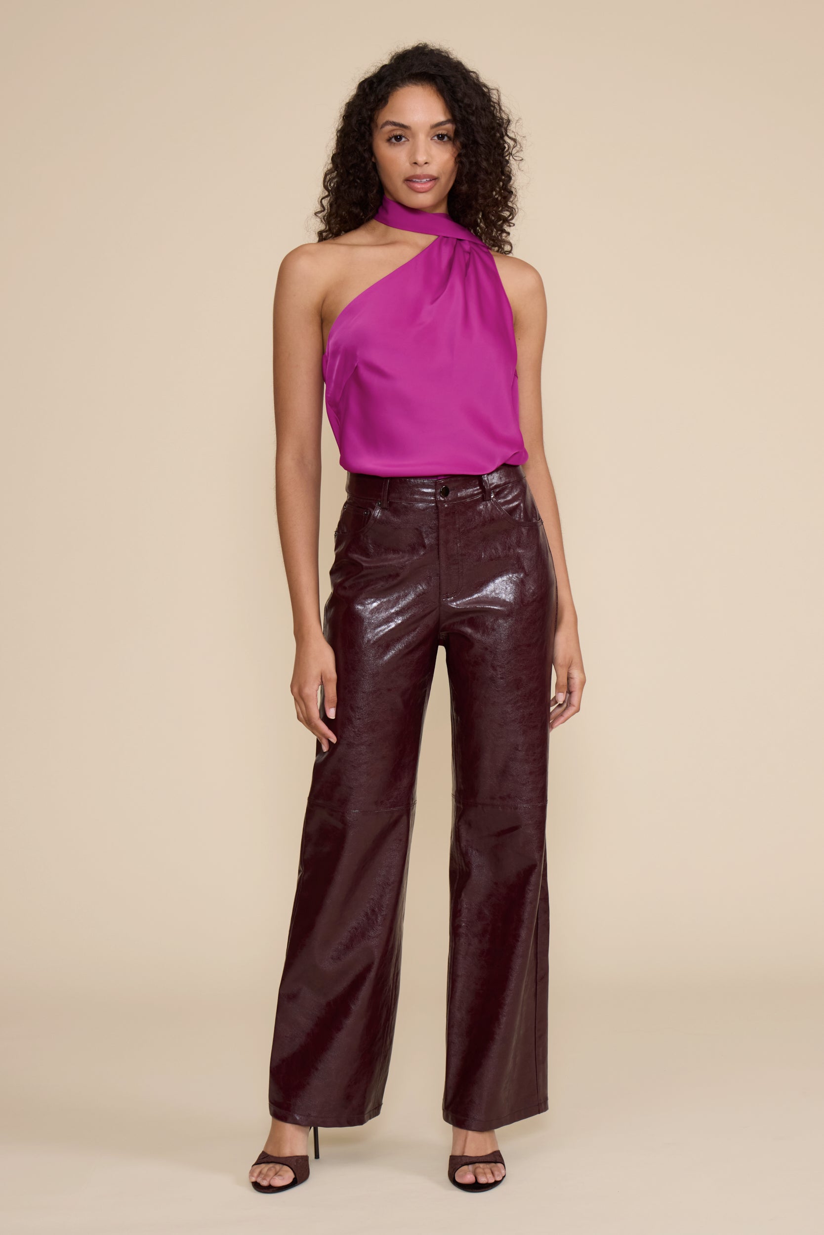 A model stands against a solid beige studio background wearing a bright magenta halter-neck top and shiny dark burgundy wide-leg pants.