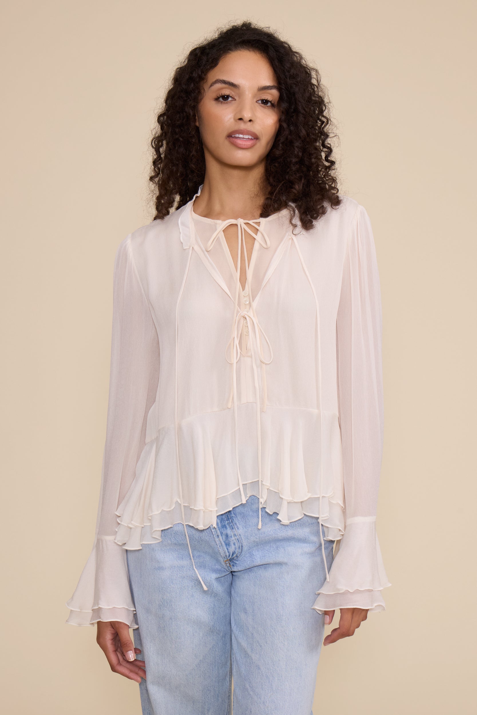 A woman modeling a sheer, long-sleeve blouse in a very light cream or off-white color with a tie-front keyhole detail and ruffled layers, paired with light-wash blue jeans against a solid beige studio background.