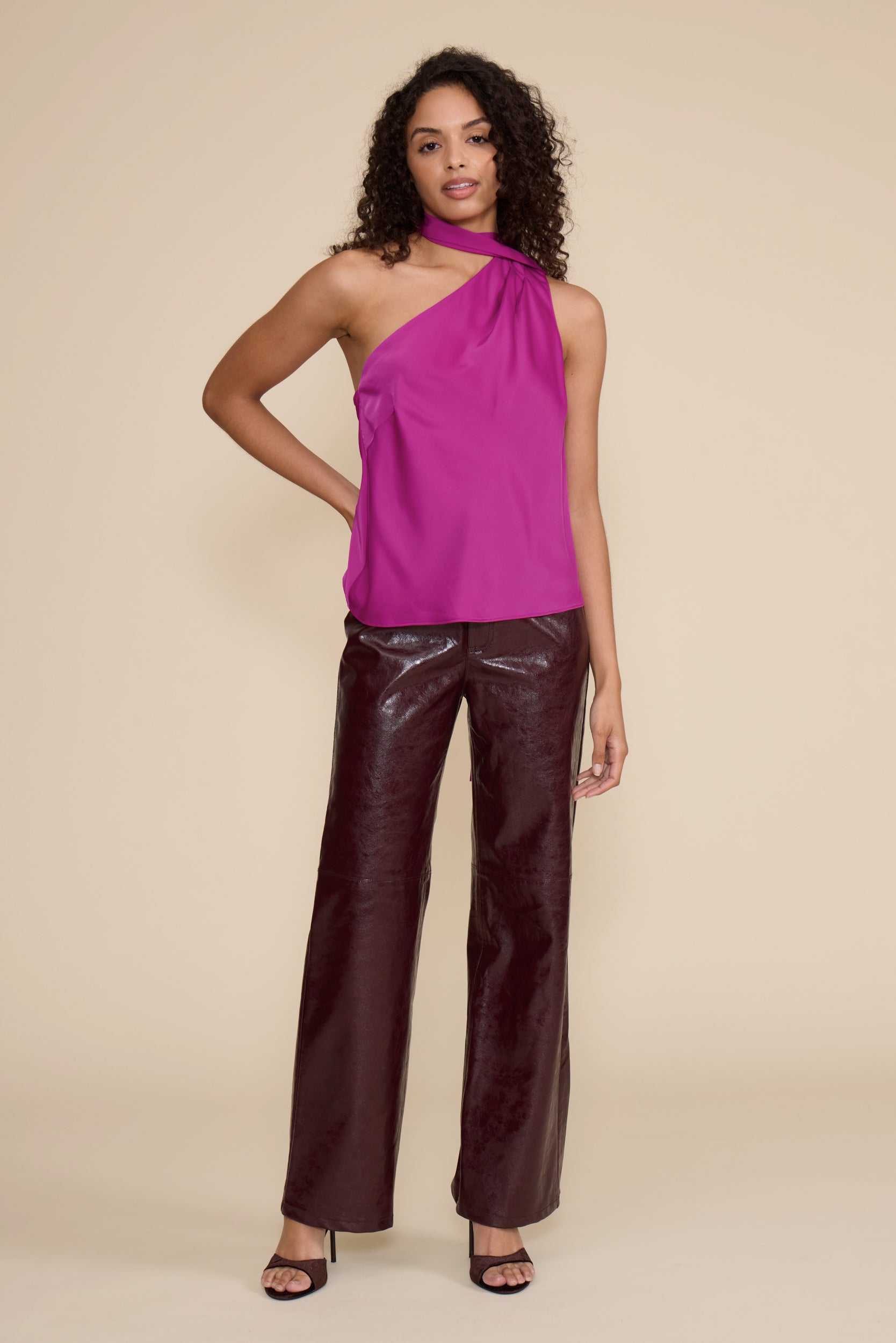 A woman modeling a bright magenta one-shoulder satin top with a high neck and dark brown patent leather pants, set against a solid beige studio background.