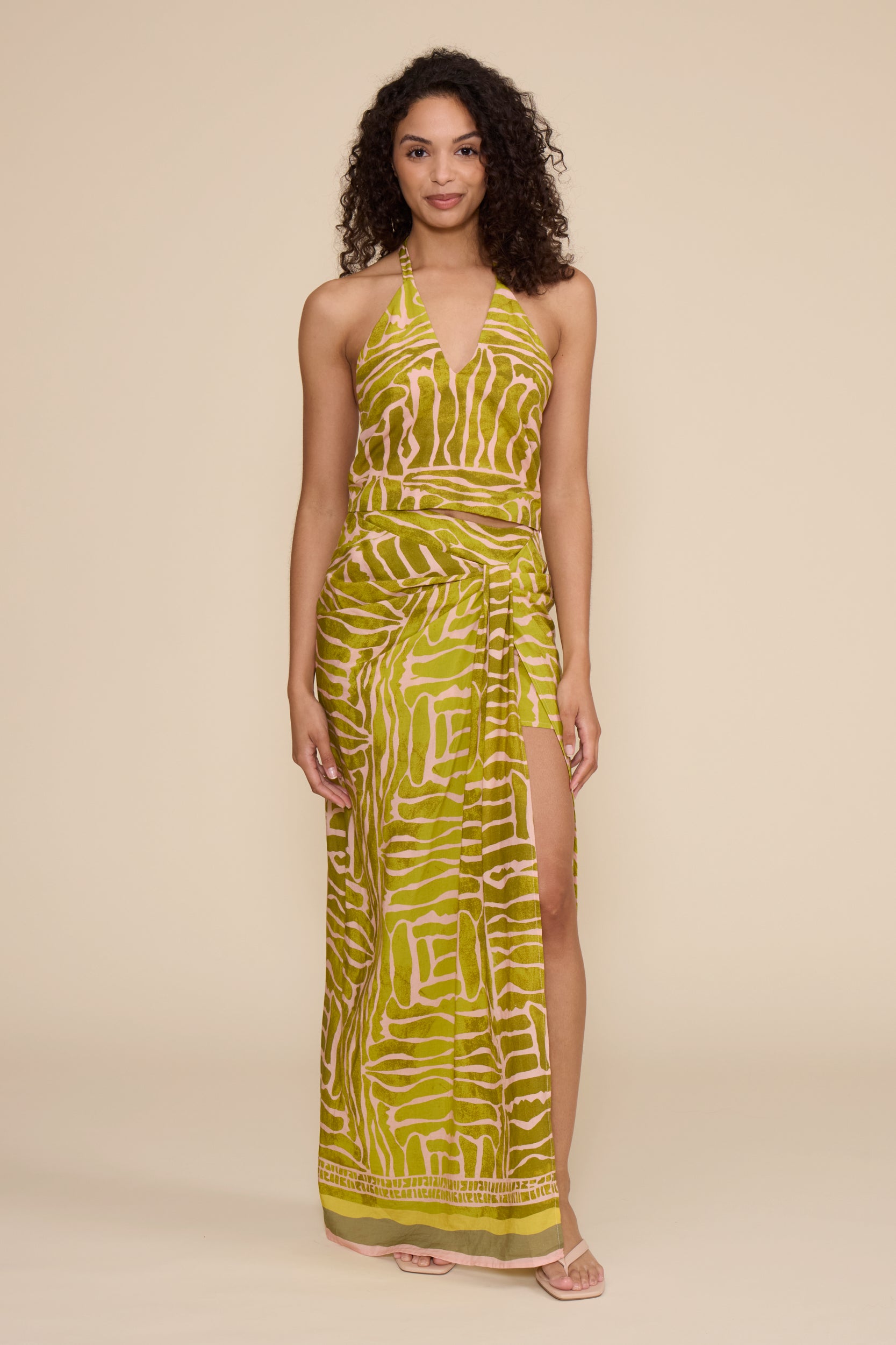 A woman shown from the back wearing a sleeveless halter-neck maxi dress with an abstract wavy print in olive green and cream, set against a solid beige studio background.