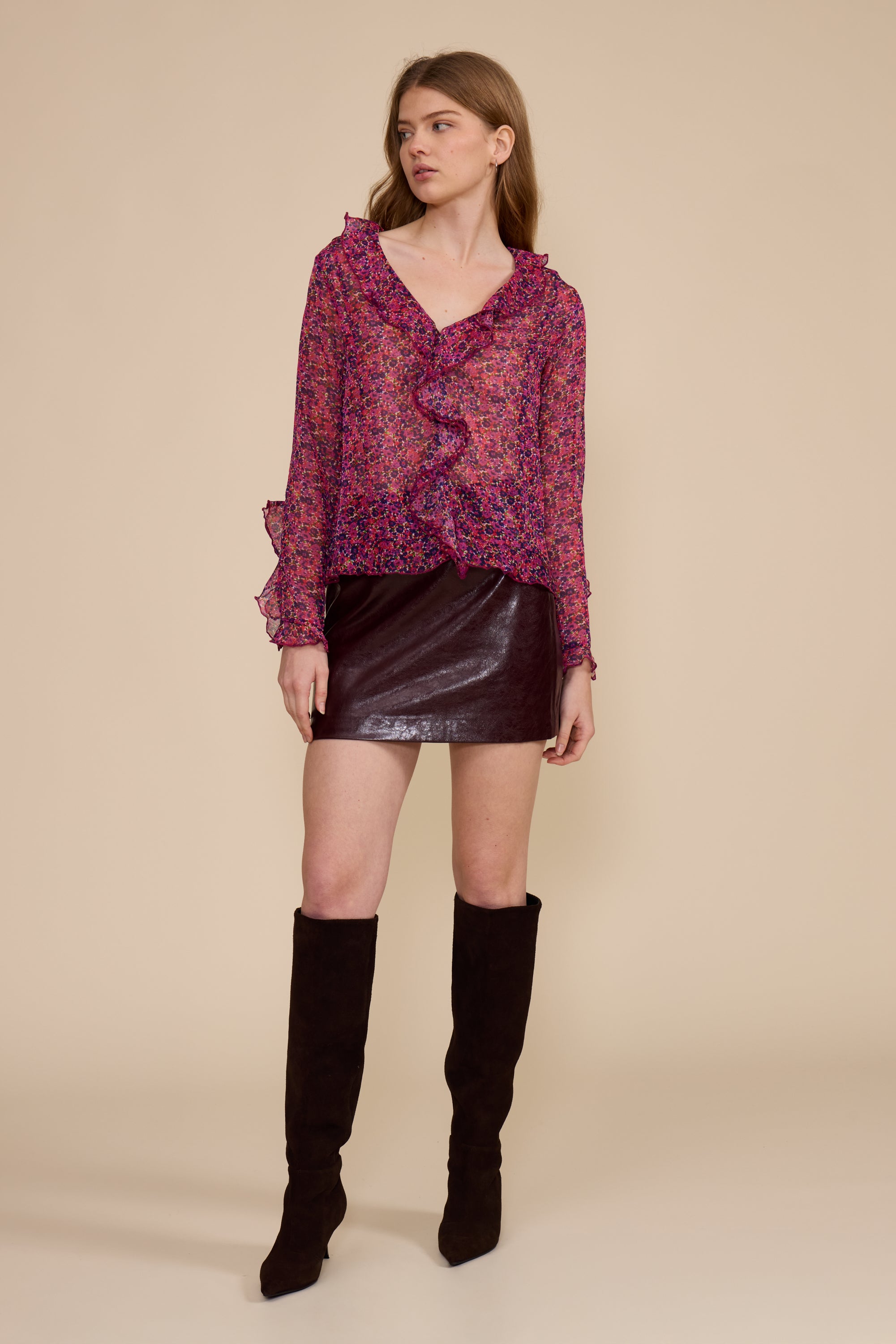 A model stands against a solid beige studio background wearing a long-sleeve ruffled blouse with a pink and purple floral pattern, a dark brown faux-leather mini skirt, and black knee-high boots.