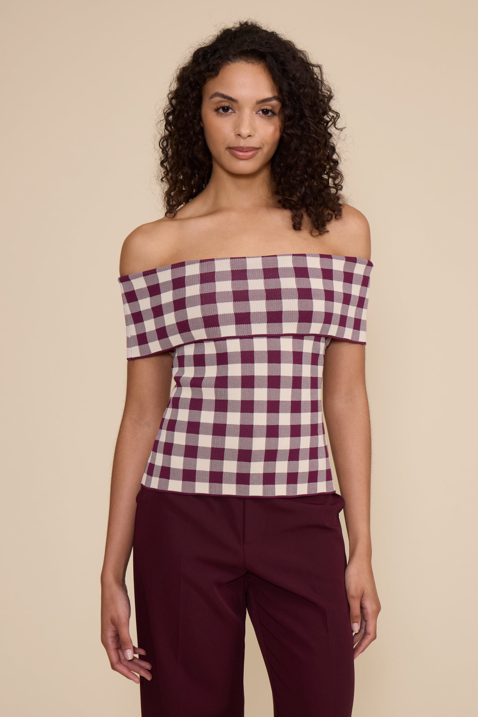 A woman modeling an off-the-shoulder gingham top in burgundy and cream, paired with burgundy pants, against a solid beige background.