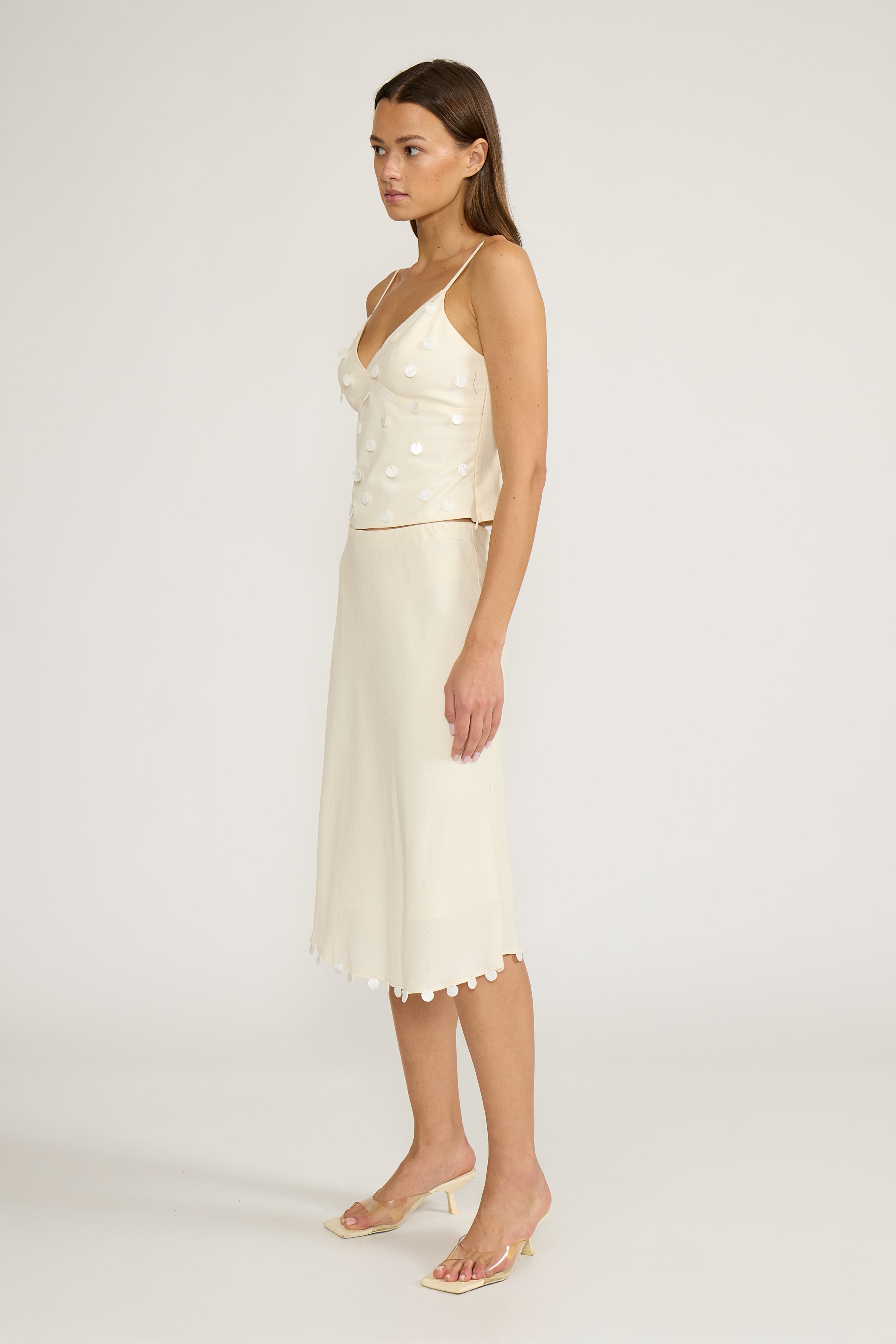 A woman viewed from the back wearing a light cream spaghetti-strap dress with a structured bodice and a flared skirt with a scalloped edge, paired with beige pointed-toe heels against a plain light gray studio background.