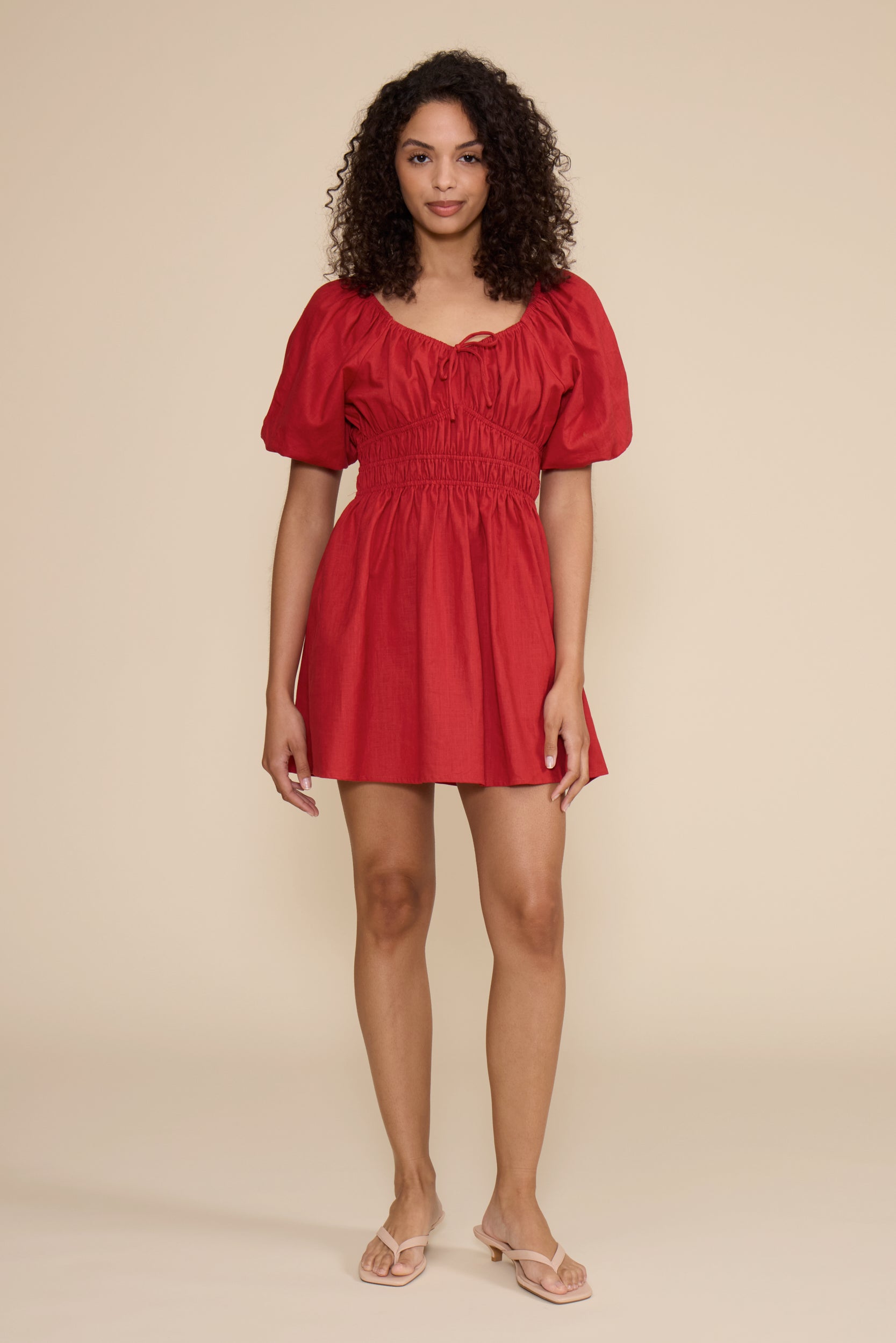 A woman modeling a short red puff-sleeve dress with a smocked waist, standing in profile against a solid beige studio background.