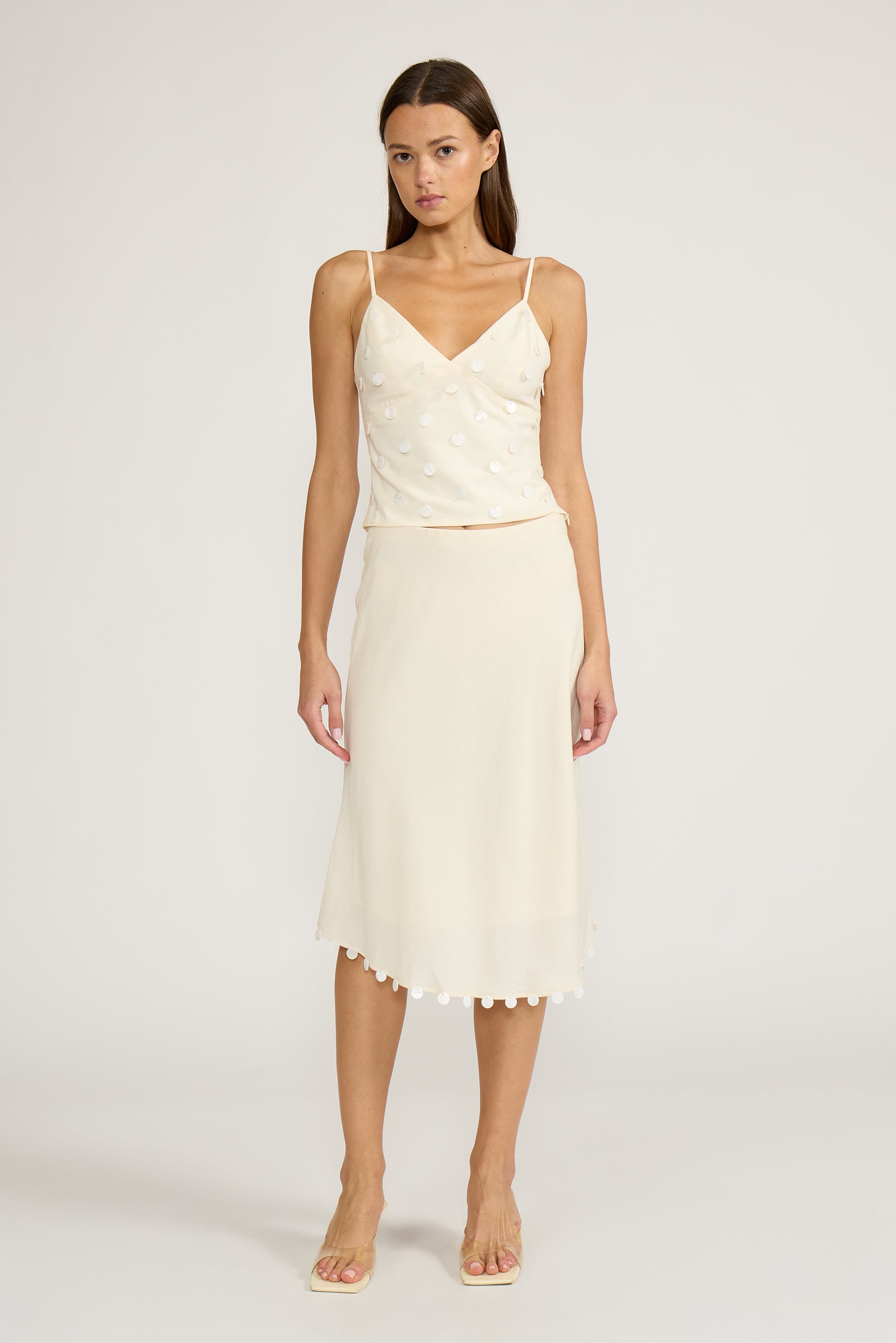 A woman modeling a two-piece cream or off-white camisole and midi skirt set against a plain light gray studio background.