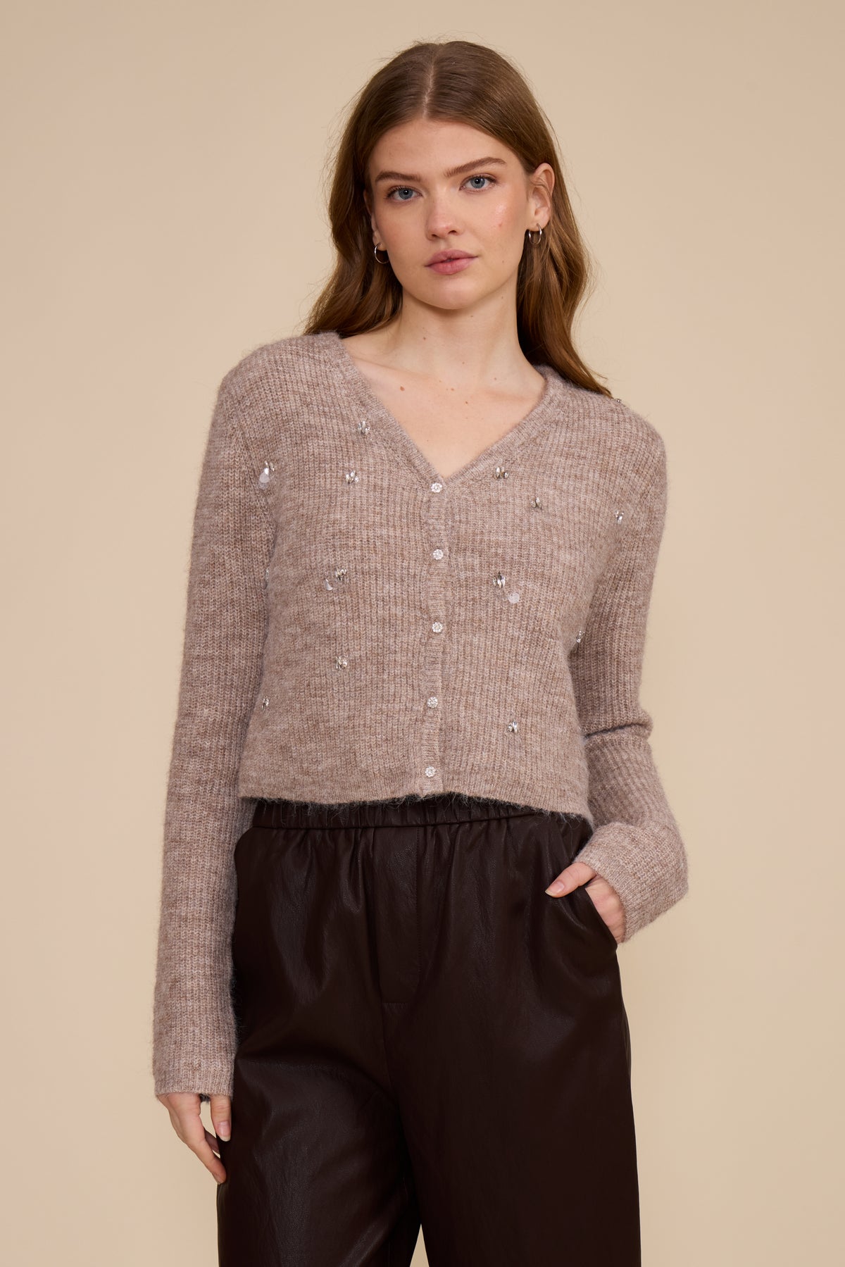 A woman shown from the back wearing a textured, heathered taupe knit sweater and dark brown pants, set against a solid beige studio background.