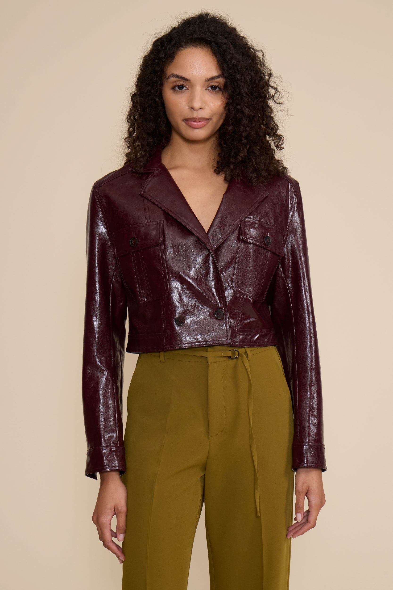 A woman modeling a cropped, glossy burgundy faux-leather jacket with flap chest pockets, paired with olive green trousers, against a solid beige background.