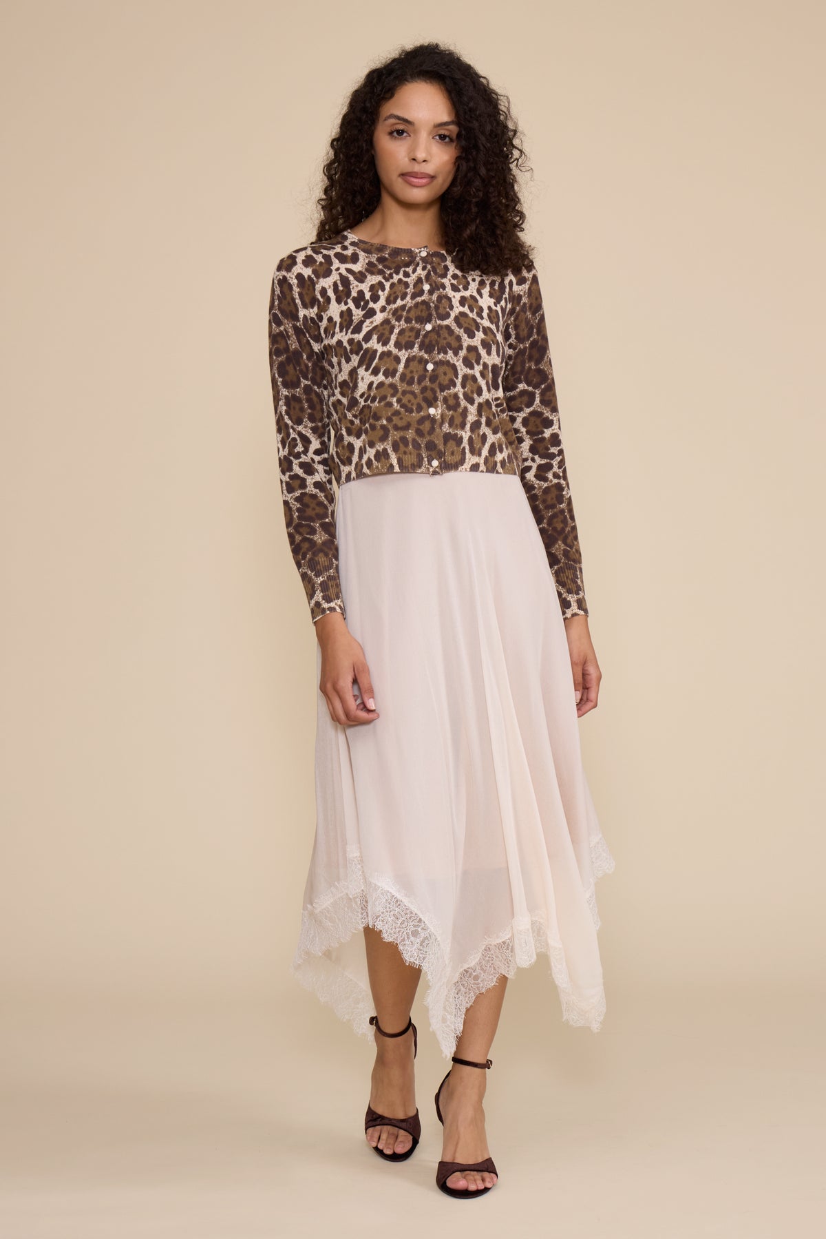A model stands against a solid beige studio background wearing a long-sleeve leopard-print top and a white high-low skirt with lace trim, paired with black strappy heels.