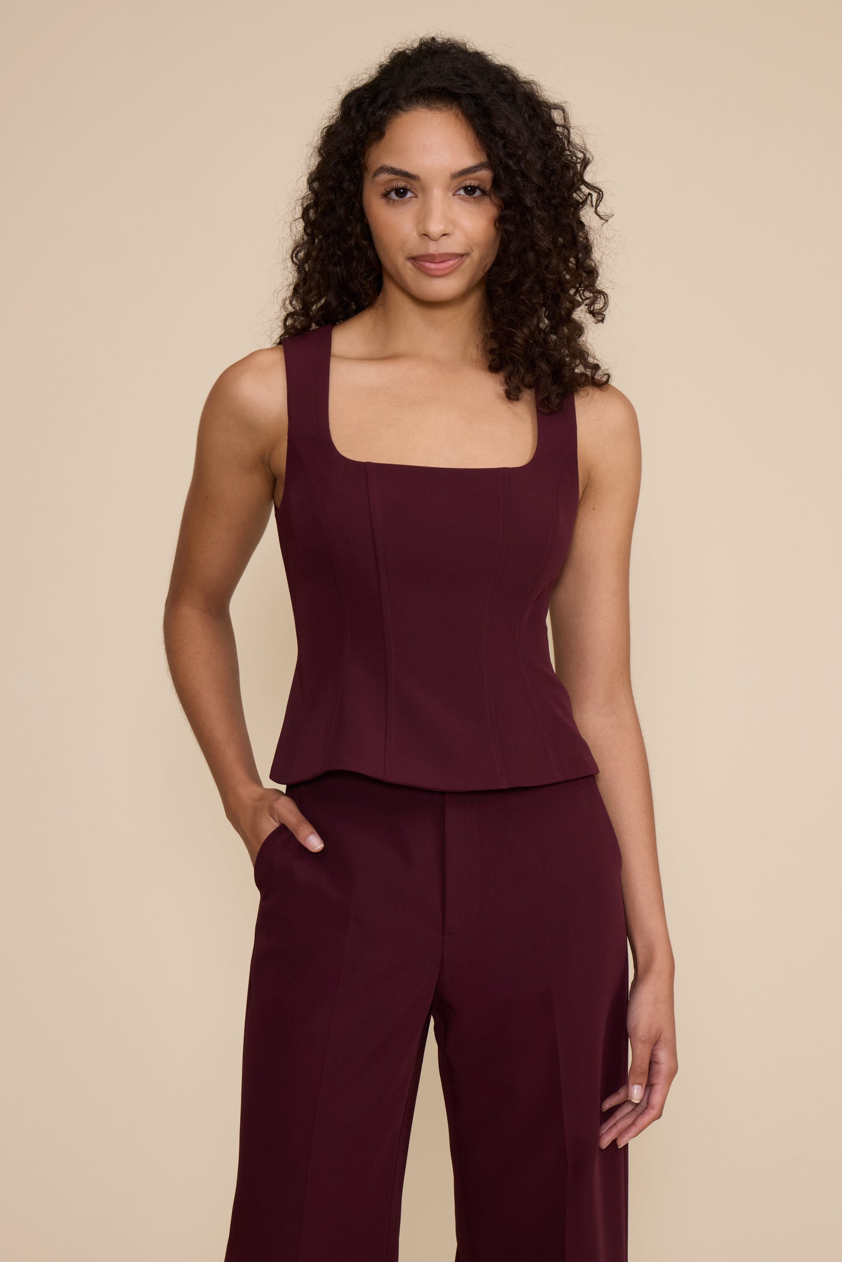 A woman modeling a sleeveless burgundy top with a square neckline and structured bodice, paired with matching wide-leg trousers, against a solid beige background.