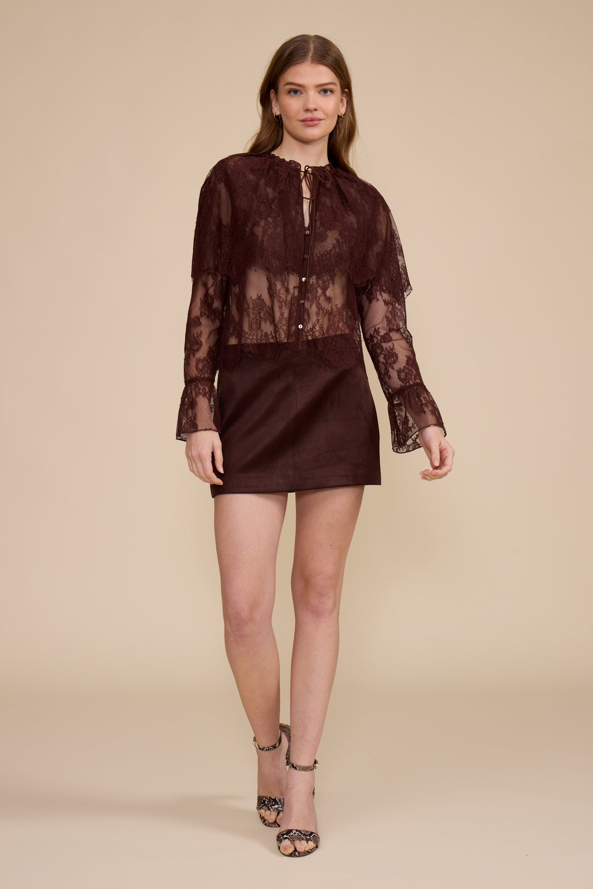 A model wearing a dark brown sheer lace long-sleeve top with button details over a matching dark brown satin mini skirt, paired with patterned high-heeled sandals, set against a solid beige studio background.