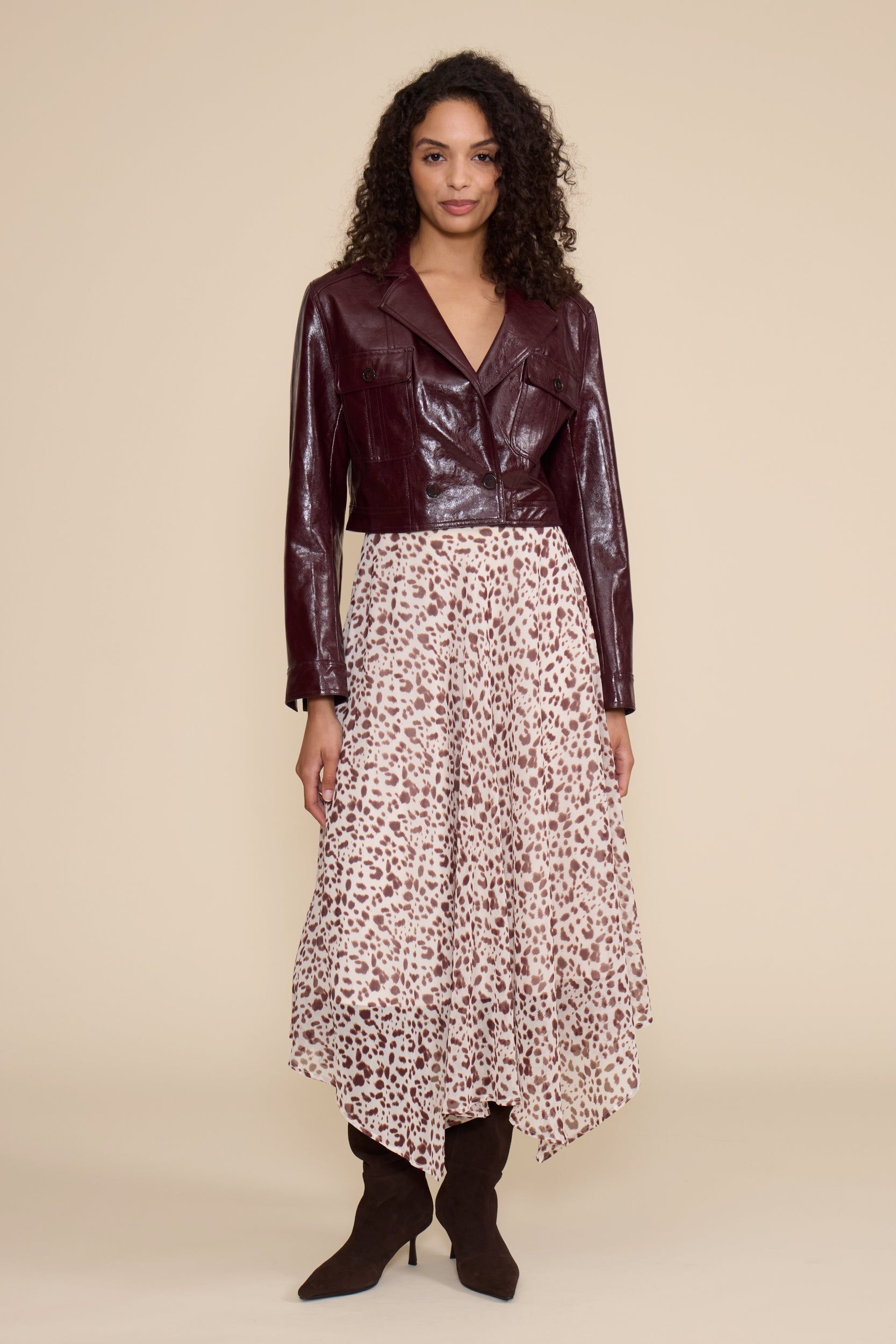 A model stands against a solid beige studio background wearing a dark burgundy faux-leather cropped jacket, a high-waisted midi skirt with a brown and beige leopard print, and dark brown pointed-toe heeled boots.