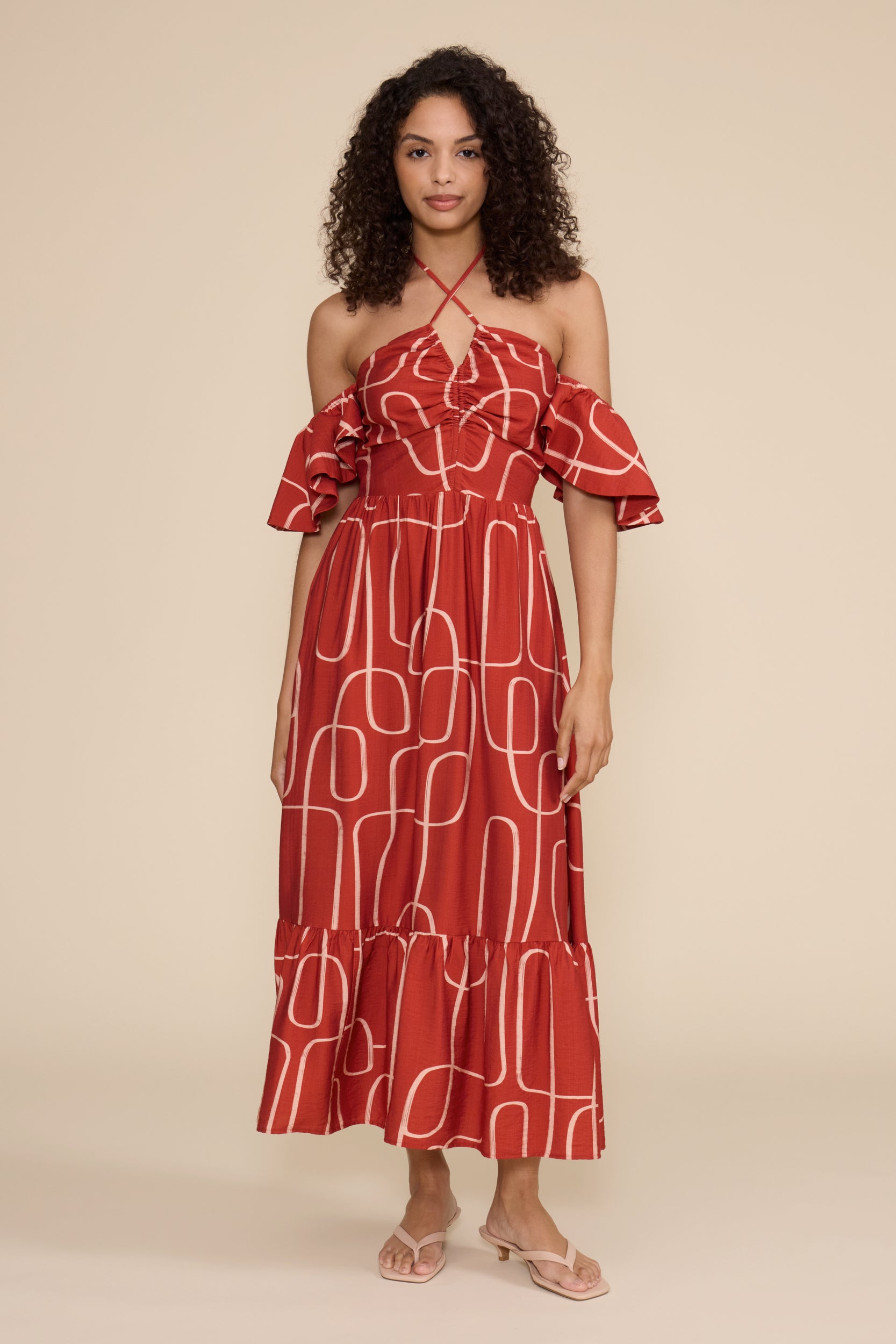 A woman modeling an off-the-shoulder red maxi dress with a white geometric line pattern, paired with light pink heeled sandals, against a solid beige studio background.
