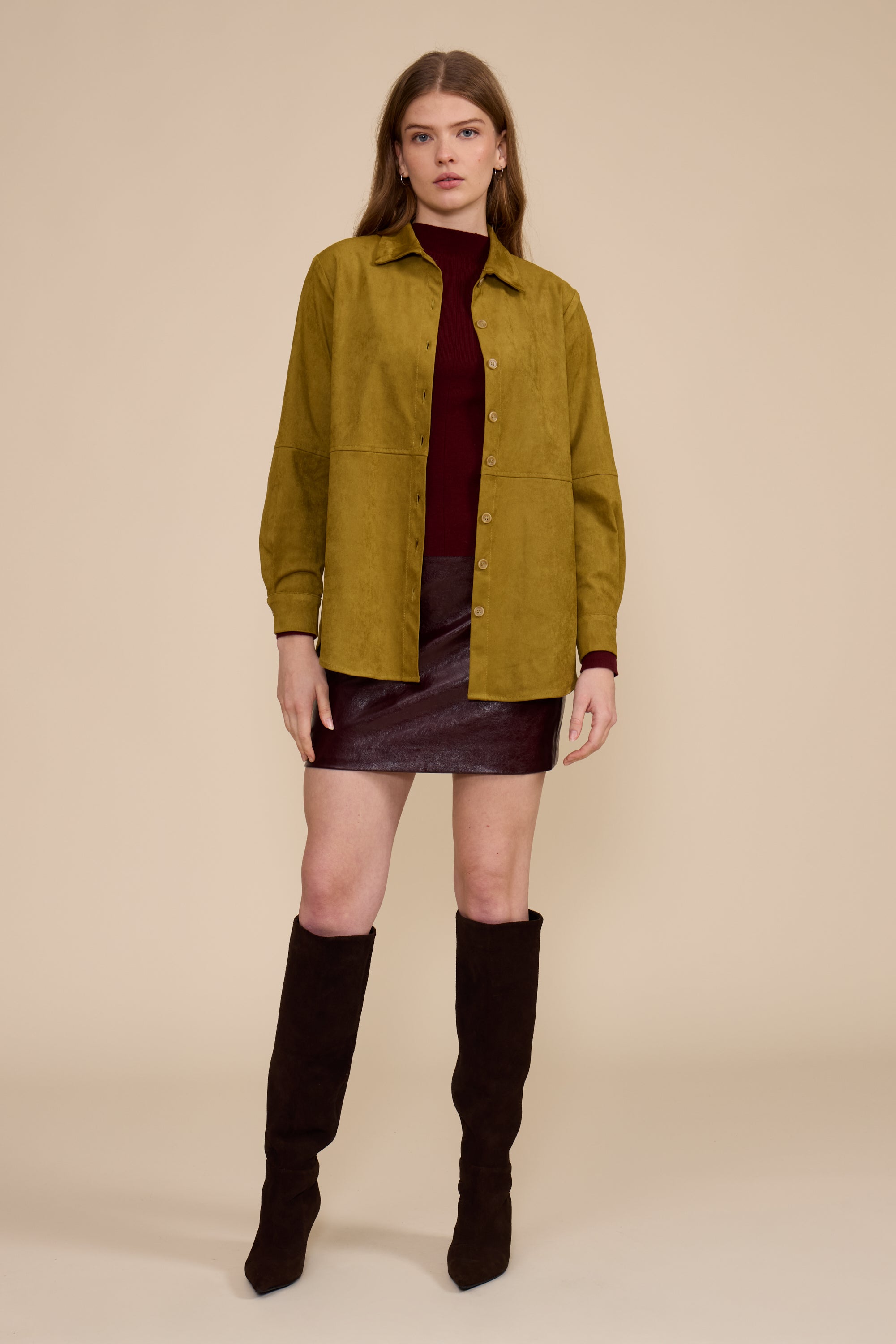 A model stands against a solid beige studio background wearing an olive green button-up overshirt over a dark burgundy turtleneck, a dark burgundy mini skirt, and dark brown knee-high boots.