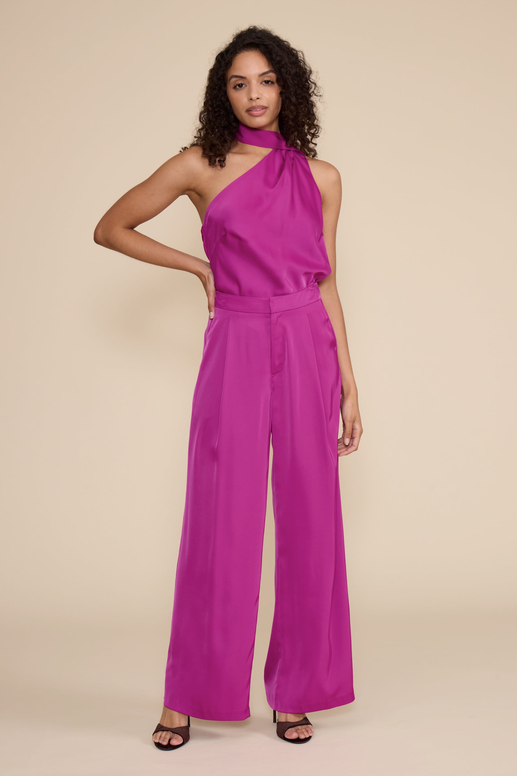 A woman modeling a bright magenta halter-neck jumpsuit with wide legs, standing against a solid beige studio background.