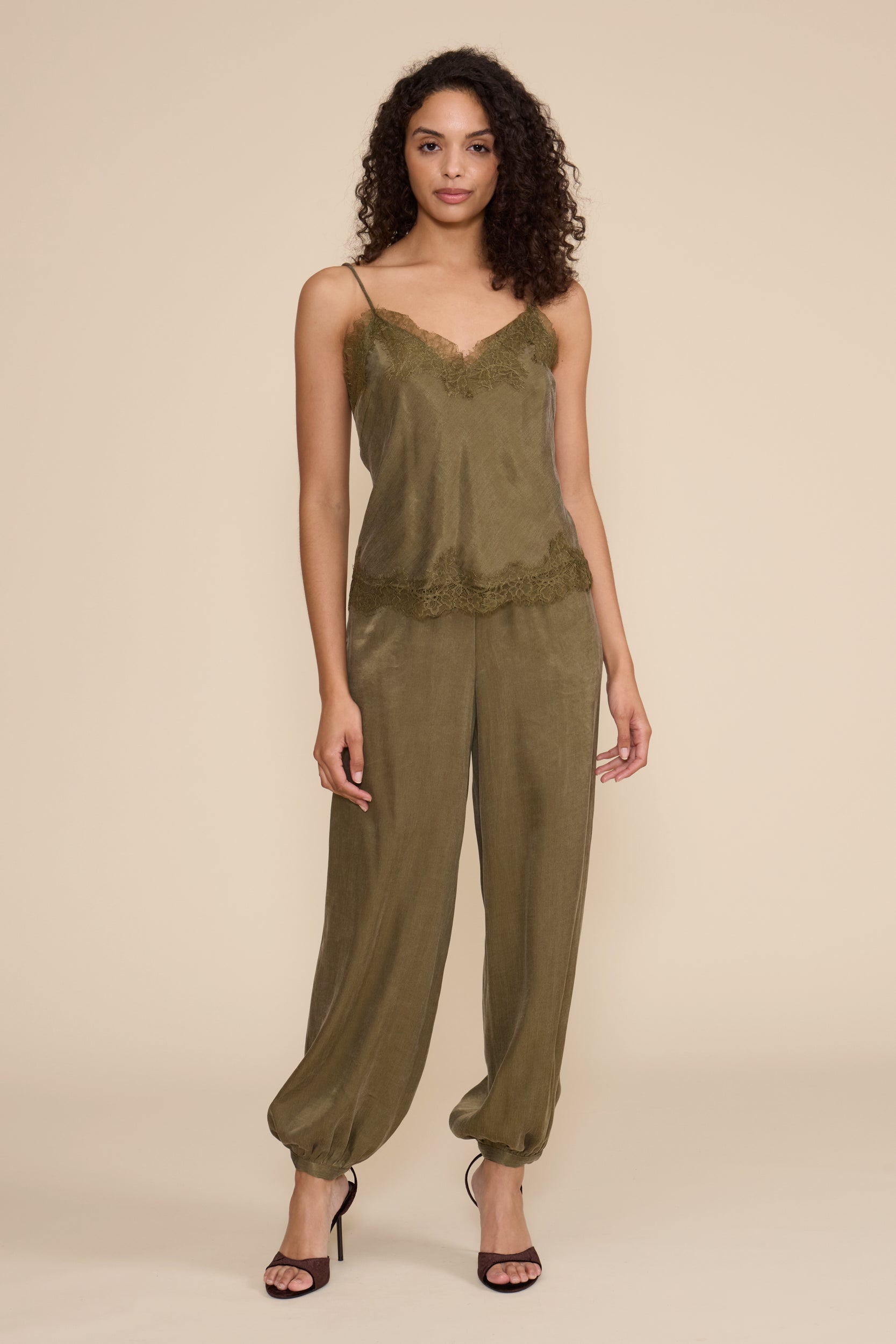 A woman modeling a sage green satin camisole and wide-leg jumpsuit set against a solid beige studio background.
