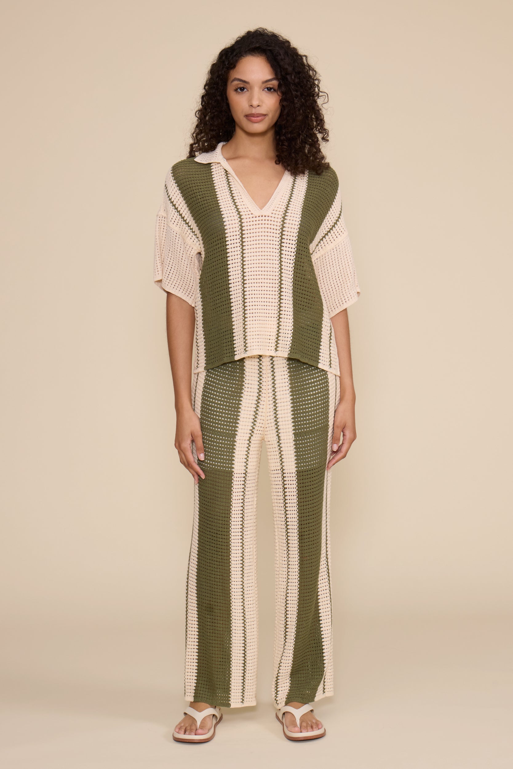 A woman modeling a matching two-piece knit set featuring a short-sleeve collared top and wide-leg pants, both in vertical stripes of olive green and cream, set against a solid beige studio background.
