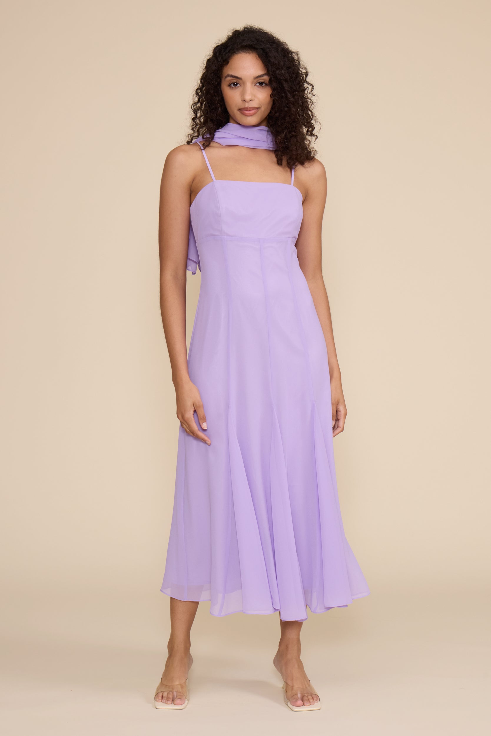 A woman modeling a light purple halter midi dress with thin straps and a matching fabric neck scarf, set against a solid beige studio background.