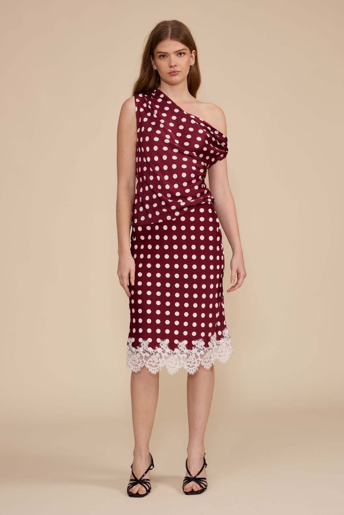 A woman modeling a one-shoulder midi dress with a burgundy and white polka dot pattern, featuring a white lace hem and a draped sleeve, set against a solid beige background. 
