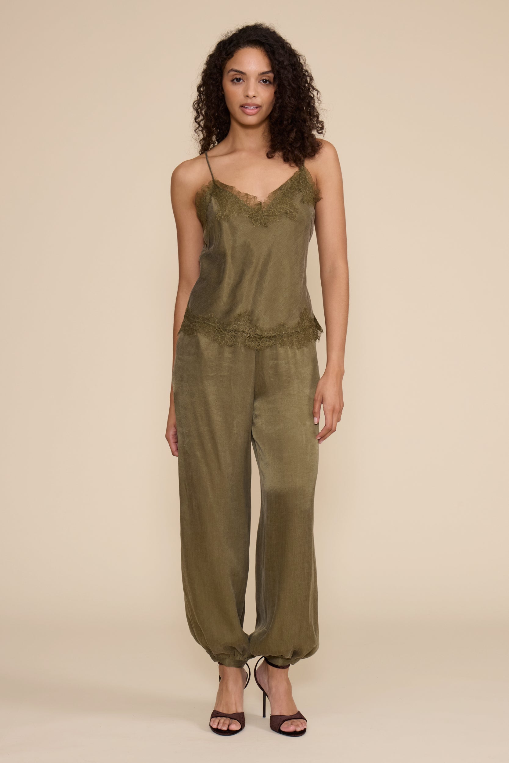 A woman modeling a sage green satin camisole and wide-leg jumpsuit set against a solid beige studio background.