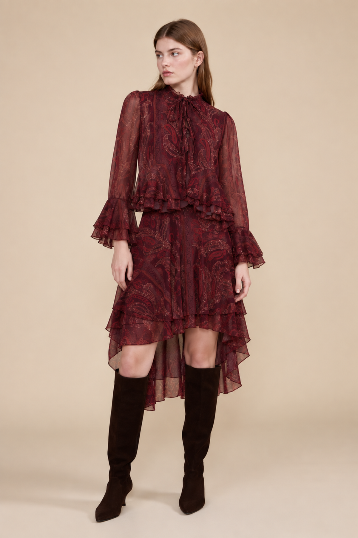 A woman wearing a sheer burgundy long-sleeve blouse with a paisley pattern, ruffled collar, and tie detail, paired with a matching skirt, set against a solid beige background.