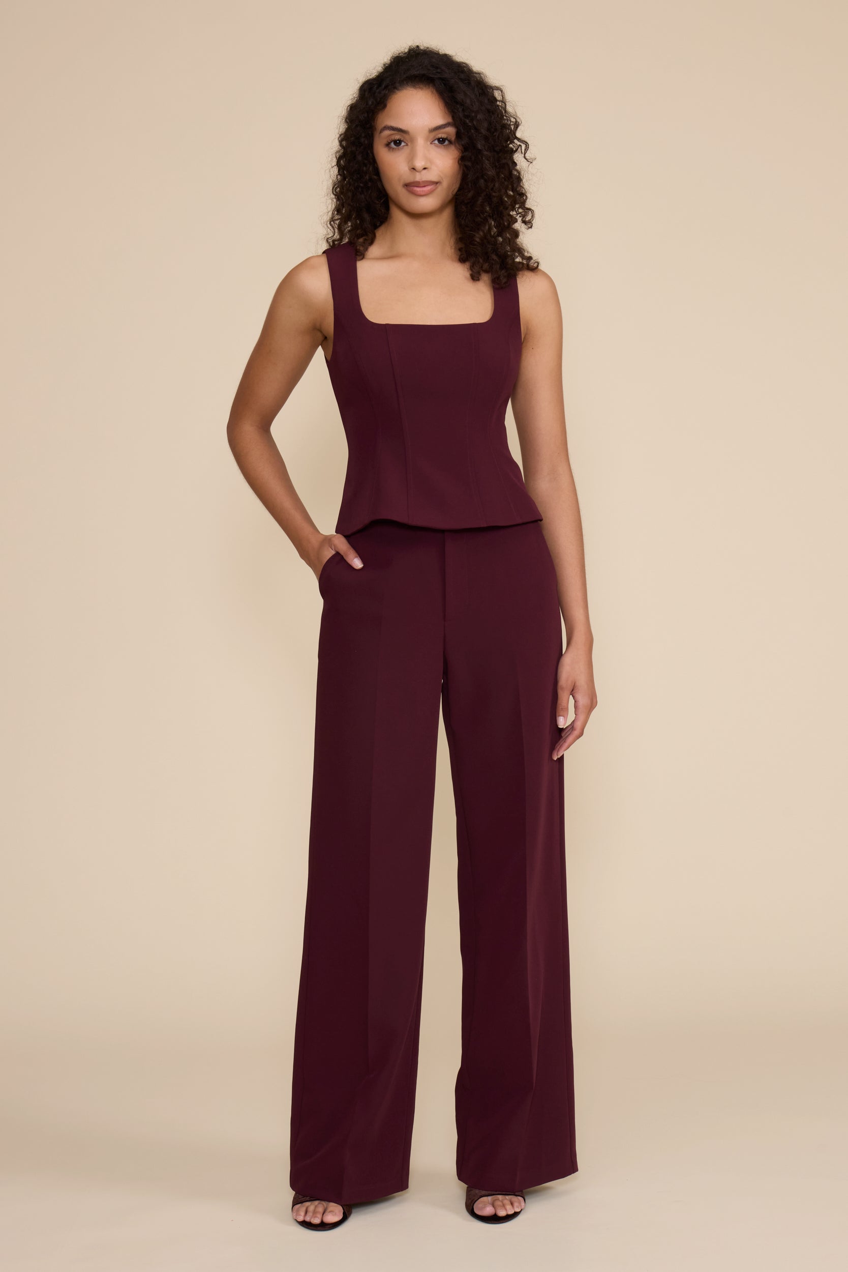 A woman modeling a sleeveless dark burgundy jumpsuit with a square neckline, peplum waist, and wide-leg pants, standing against a solid beige studio background.
