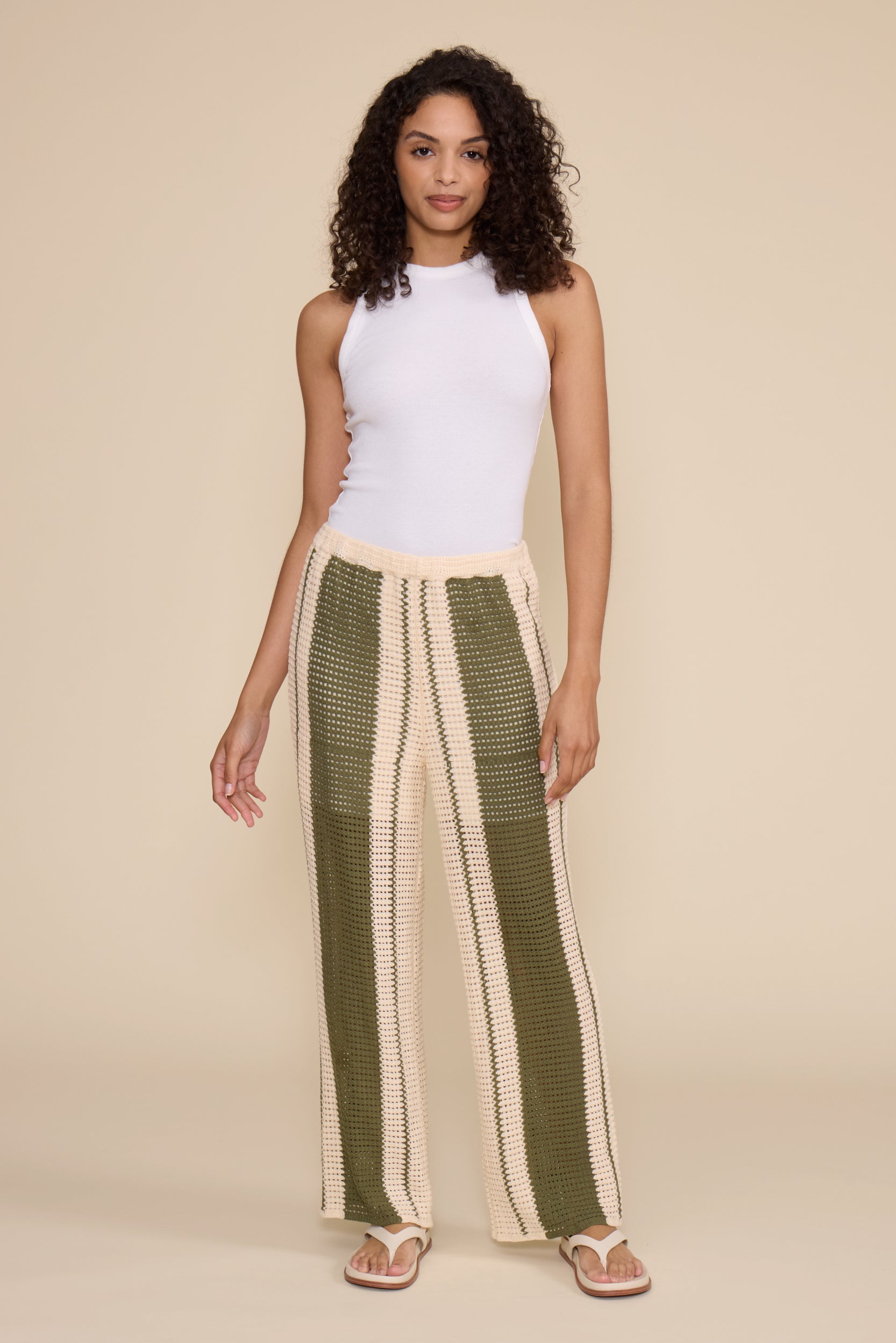 A woman modeling a matching two-piece knit set featuring a short-sleeve V-neck top and wide-leg pants in vertical olive green and cream stripes, styled with white slide sandals against a solid beige studio background.