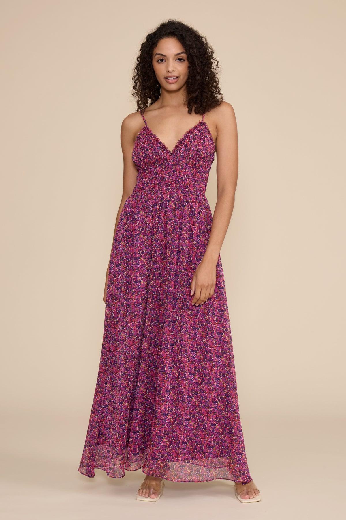 A woman modeling a sleeveless, floor-length maxi dress with thin straps and a vibrant pink, purple, and blue floral pattern against a solid beige studio background.